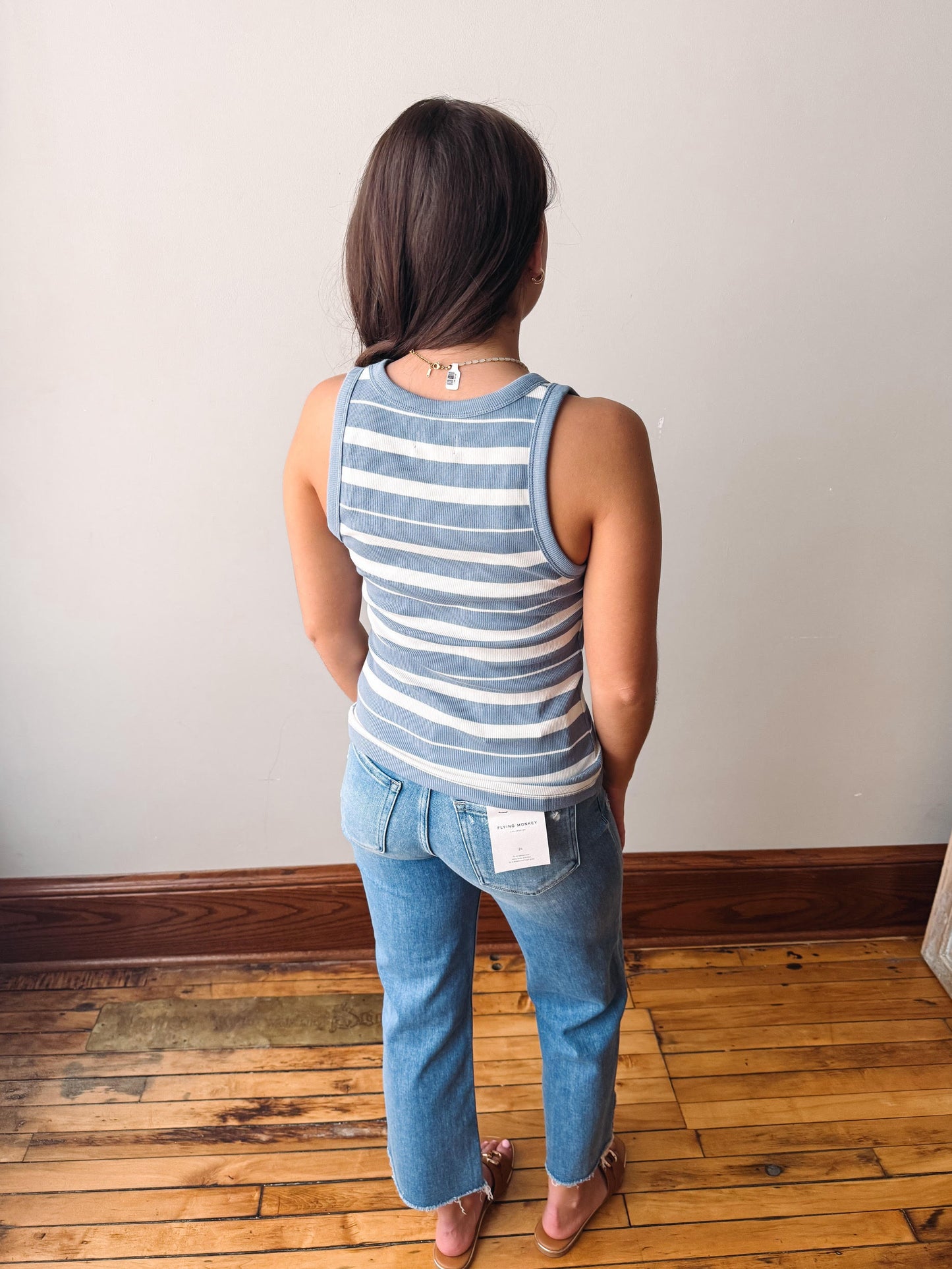Person wearing a striped tank top and jeans standing on a wooden floor with a white wall background