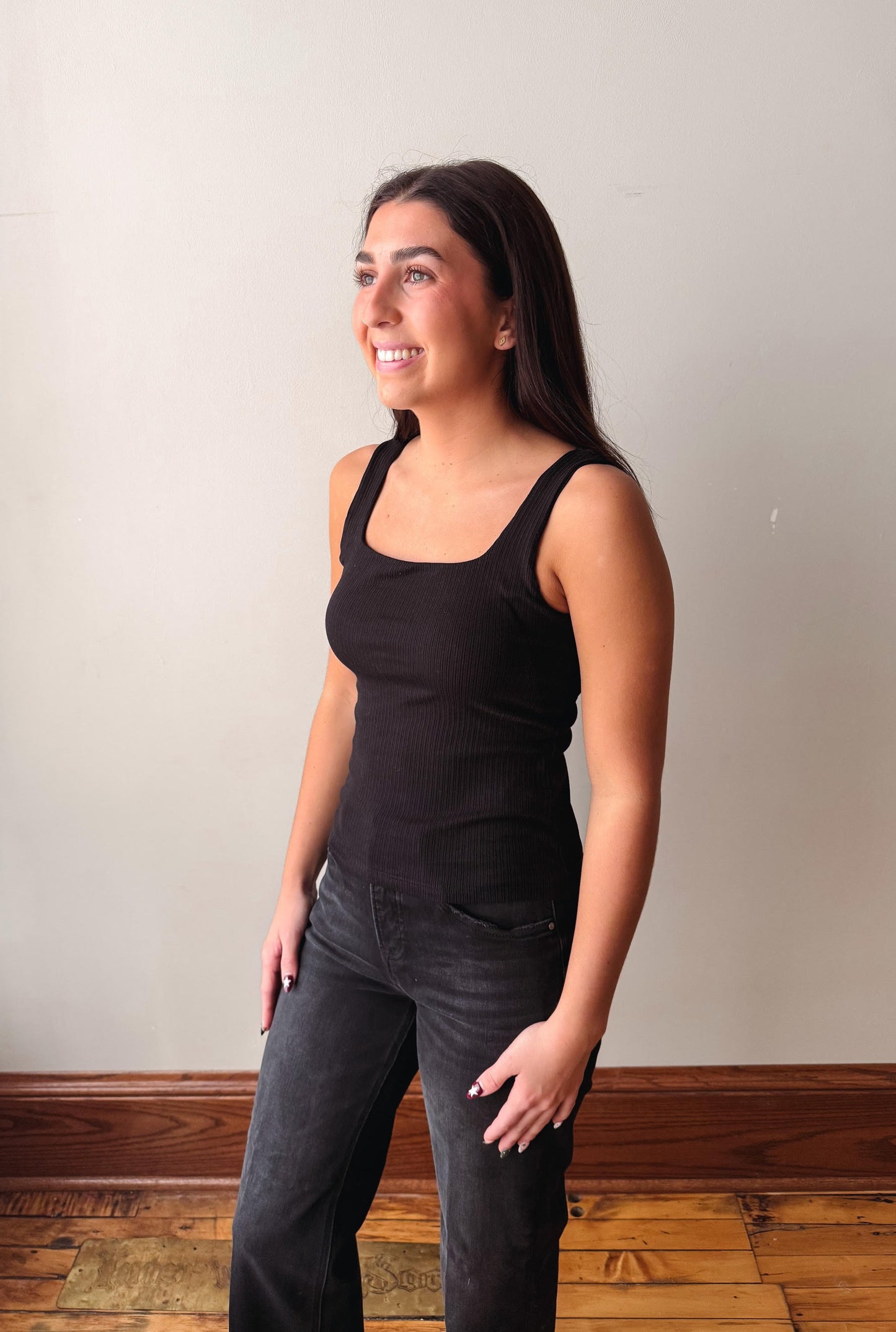 A woman wearing a black ribbed tank top and dark gray jeans, standing in profile against a plain beige wall with a wooden baseboard and hardwood floor.