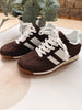 A pair of dark brown suede sneakers with white laces, white side stripes, and a white midsole, styled on a light surface with a potted plant in the background.