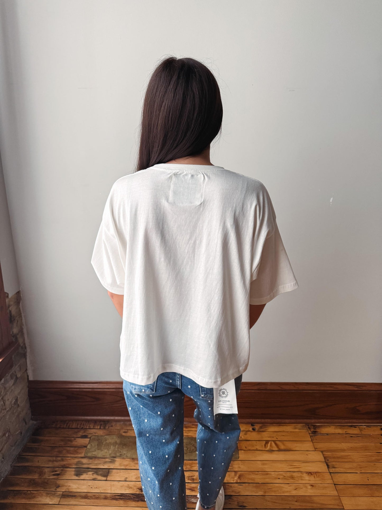 A person viewed from behind wearing an oversized off-white t-shirt and blue jeans with small white dots, standing on a wooden floor against a plain white wall.