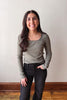 A woman with long dark hair stands indoors wearing a fitted long-sleeve top in a muted sage green color with dark charcoal jeans, posing against a plain off-white wall with a wooden baseboard and floor.