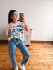 A woman takes a mirror selfie in a room with light wood floors and a white wall. She is wearing a white graphic t-shirt with blue and yellow 'Beast' lettering, blue jeans with small white polka dots, and white sneakers.