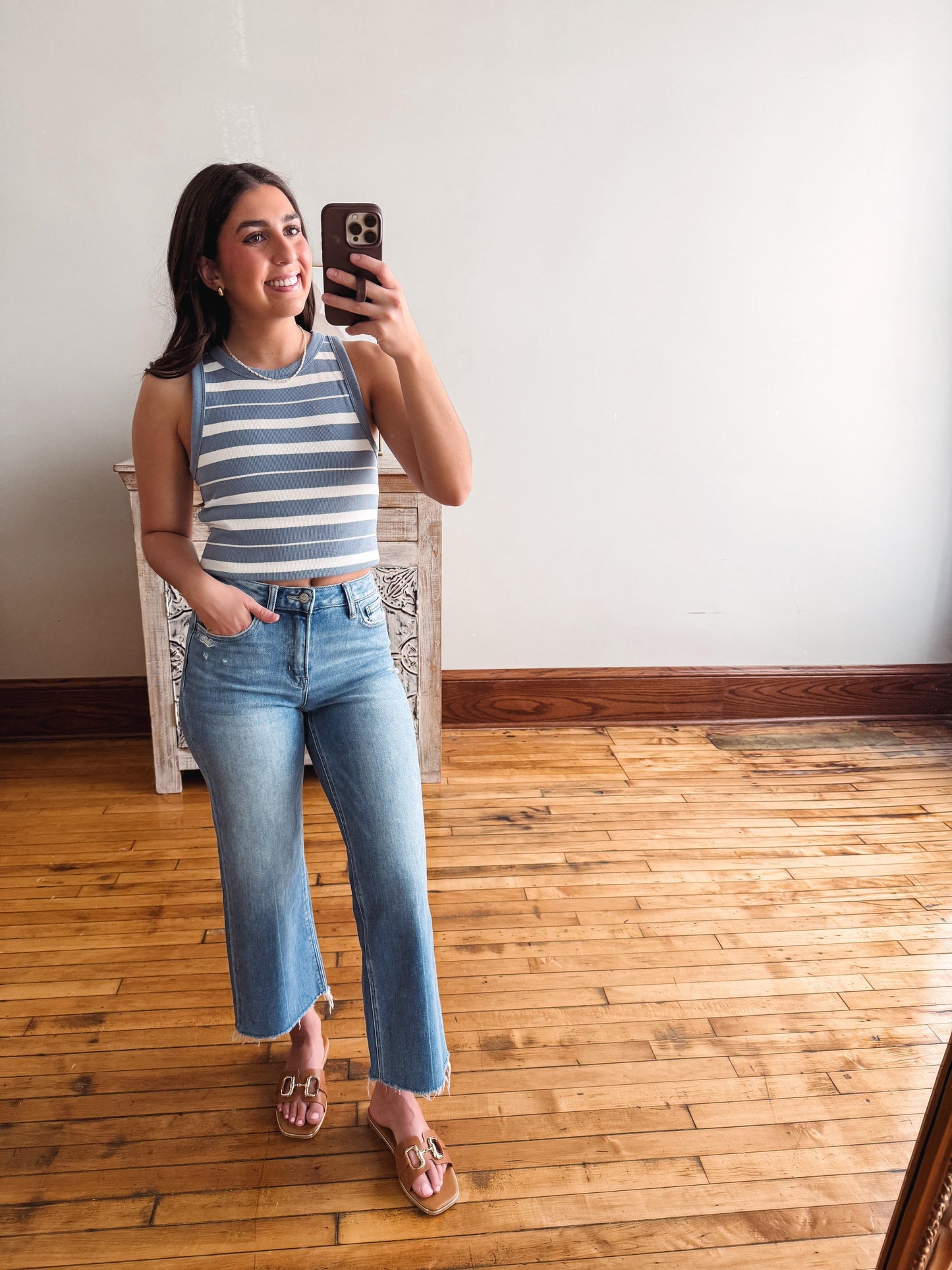 Woman taking a mirror selfie wearing a striped tank top and jeans in a room with wooden flooring.