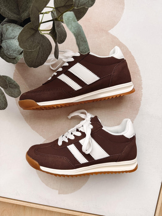 A pair of retro-style low-top sneakers in dark brown with three white stripes, white laces, white midsoles, and gum brown outsoles, arranged on a light surface with a plant leaf in the corner.