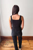 A woman viewed from behind wearing a black ribbed tank top with a square neckline and dark jeans, standing on a wooden floor against a plain white wall.