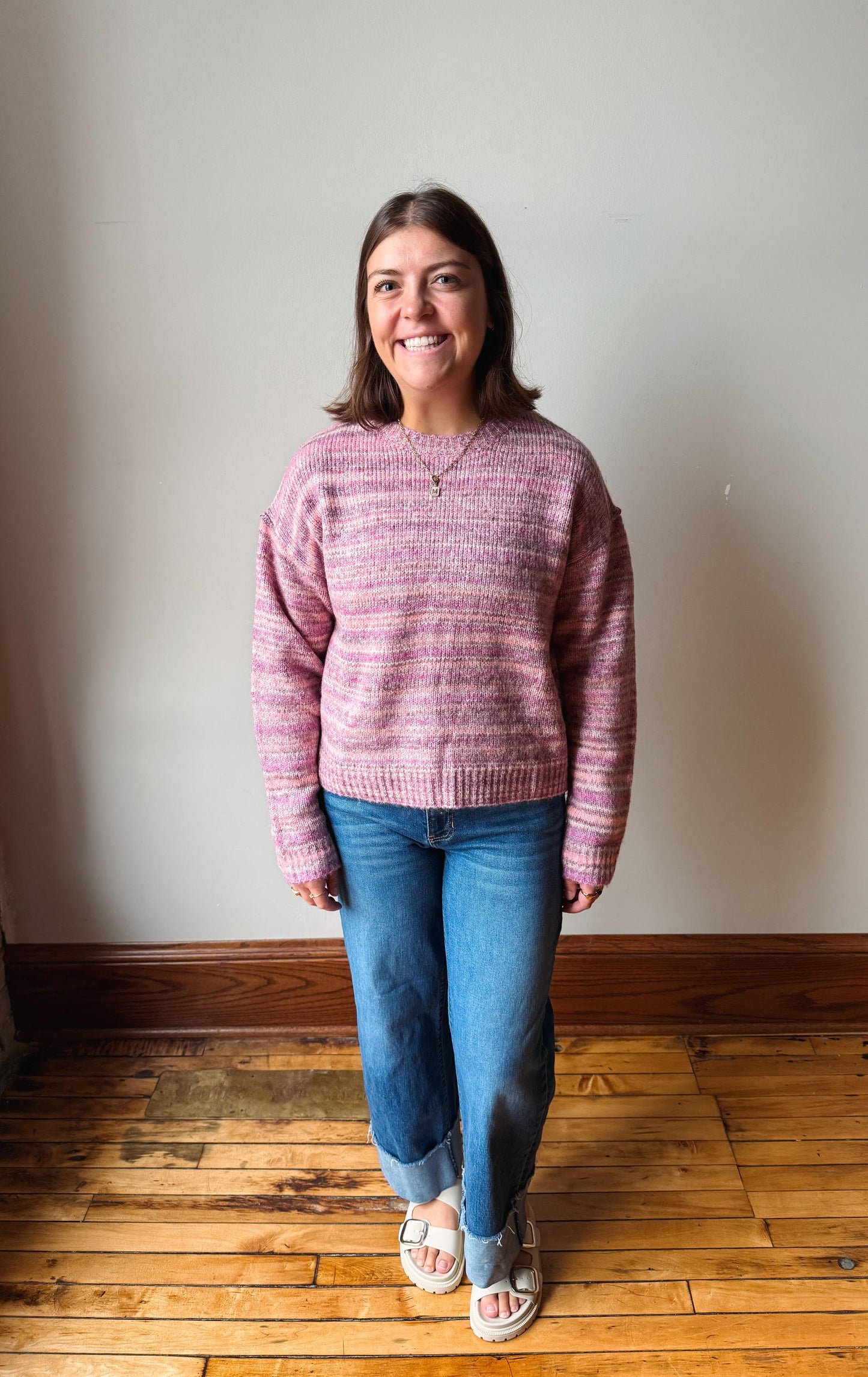 A woman standing indoors wearing a pink and purple marled knit sweater, blue jeans, and white slide sandals against a plain wall and wooden floor.