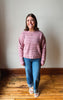 A woman standing indoors wearing a pink and purple marled knit sweater, blue jeans, and white slide sandals against a plain wall and wooden floor.