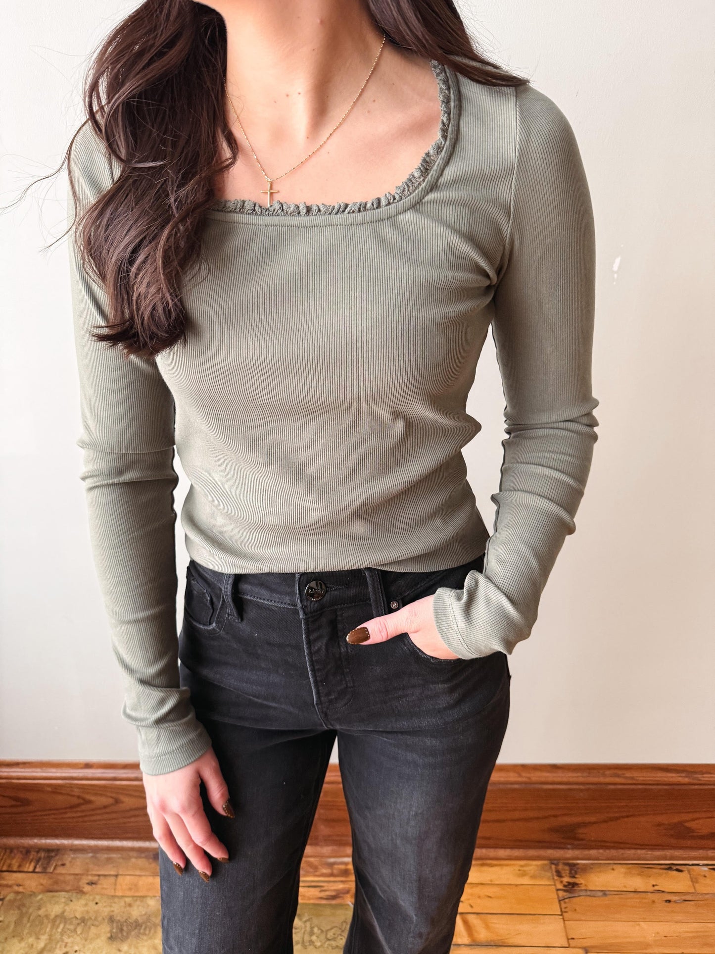 A woman wearing a ribbed long-sleeve scoop-neck top in sage green, paired with dark gray jeans, standing against a neutral indoor wall with wooden flooring.