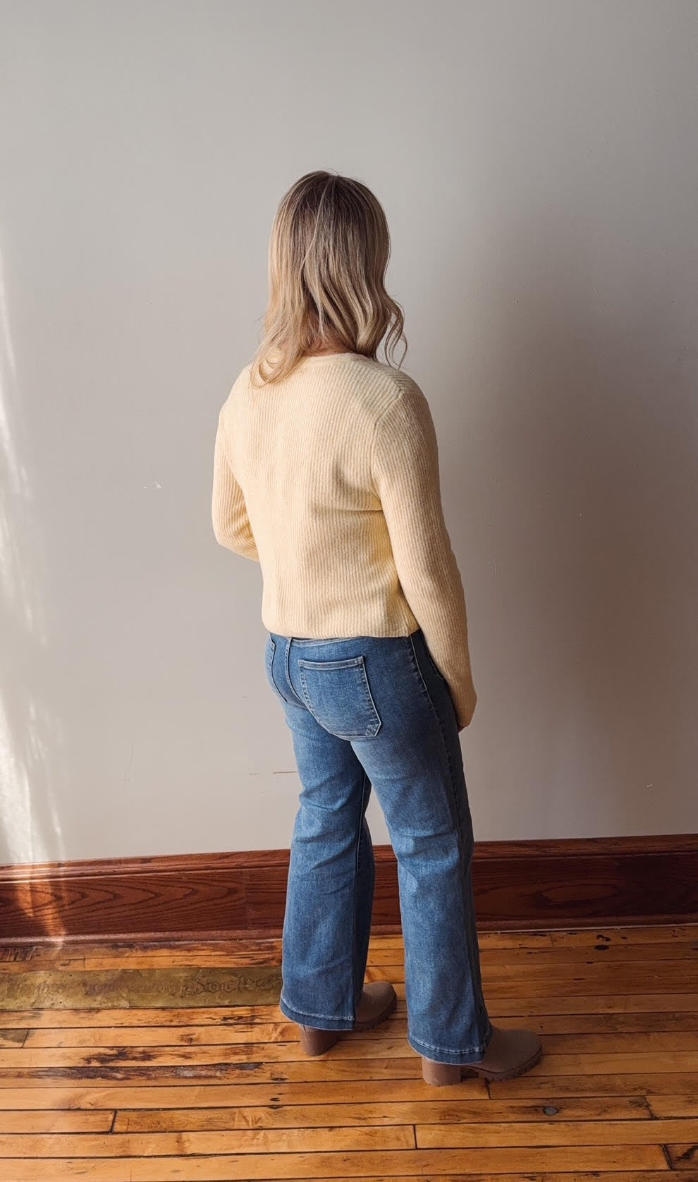 Person wearing a yellow sweater and blue jeans standing against a plain wall.