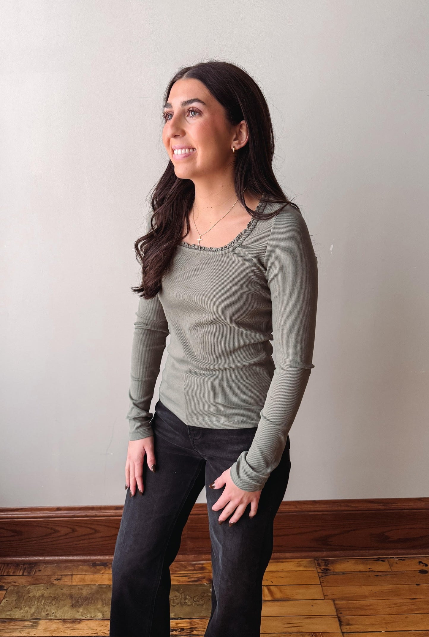 A woman with long dark hair wearing a sage green long-sleeve top and black jeans, standing in an indoor setting with a light gray wall and wooden floor.