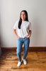 A woman stands in a room with a light gray wall and wooden floor, wearing a white short-sleeve t-shirt, blue jeans with small white star-like prints, and white sneakers.