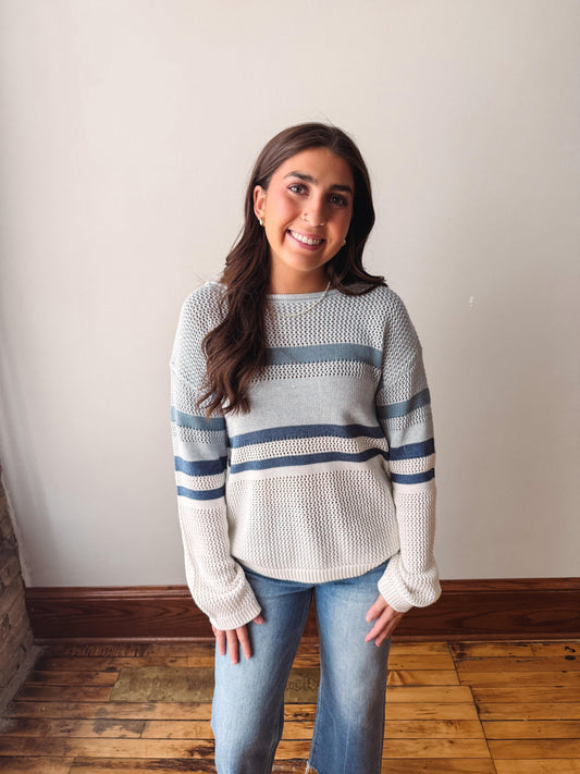 Woman wearing a light gray sweater with blue stripes against a plain wall.