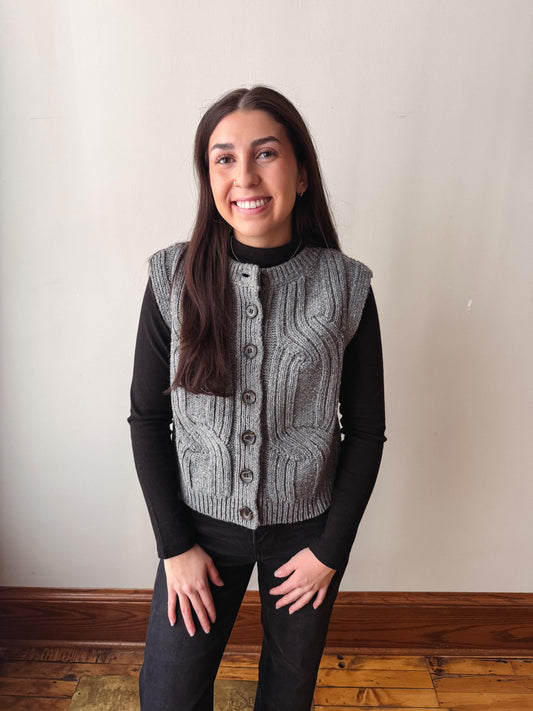 A woman wearing a gray cable-knit button-front vest over a black long-sleeve top and black pants, standing against a plain off-white wall with wood flooring visible.