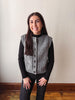 A woman wearing a gray cable-knit button-front vest over a black long-sleeve top and black pants, standing against a plain off-white wall with wood flooring visible.
