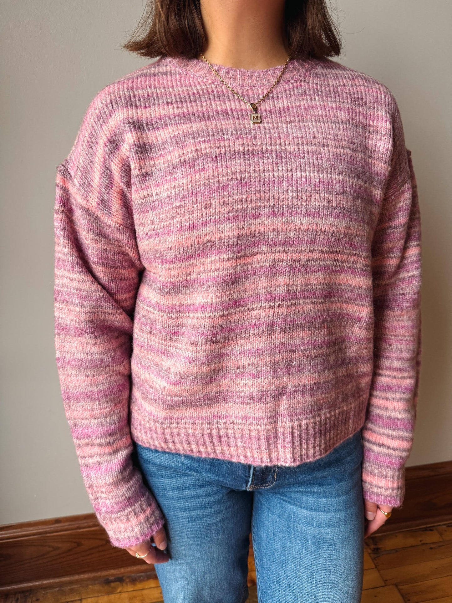 A person wearing a pink and purple marled knit sweater with blue jeans, standing indoors against a plain wall.