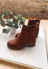 A pair of brown suede ankle boots with elastic side gore panels and stacked block heels, displayed on a white board with a potted eucalyptus plant and a brick wall background.