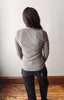 A woman viewed from behind wearing a light gray long-sleeve knit top and dark jeans, standing on a wooden floor against a plain off-white wall.