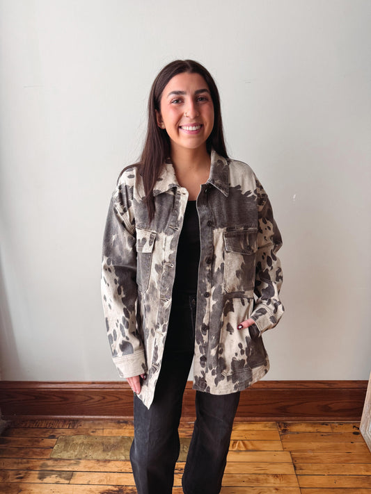 A woman stands indoors wearing a loose, oversized camouflage-patterned jacket over a black top and black pants, against a plain off-white wall with a wooden floor.