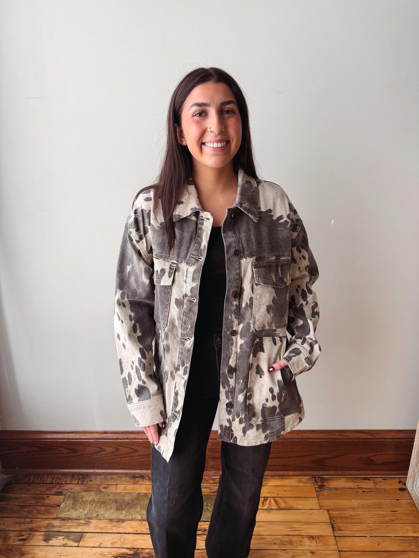 A woman stands indoors wearing a loose, oversized camouflage-patterned jacket over a black top and black pants, against a plain off-white wall with a wooden floor.