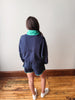 Person wearing a navy blue outfit with green accents standing on a wooden floor against a white wall.