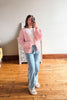A woman takes a mirror selfie in a room with hardwood floors, wearing a pink knit cardigan over a white t-shirt, light blue jeans, and white slide sandals.
