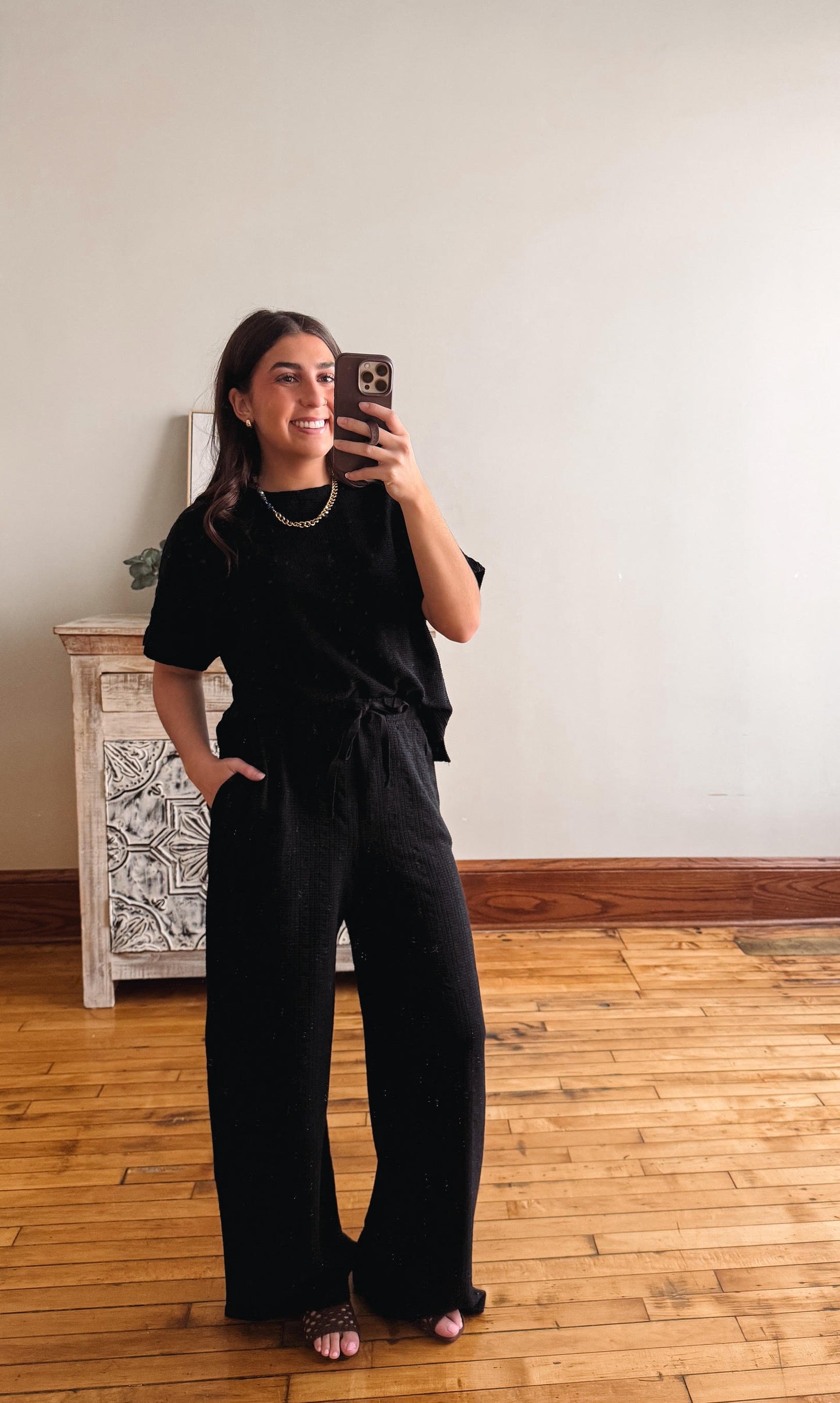 Woman taking a mirror selfie wearing a black outfit in a room with wooden flooring and a white wall.