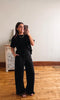 Woman taking a mirror selfie wearing a black outfit in a room with wooden flooring and a white wall.