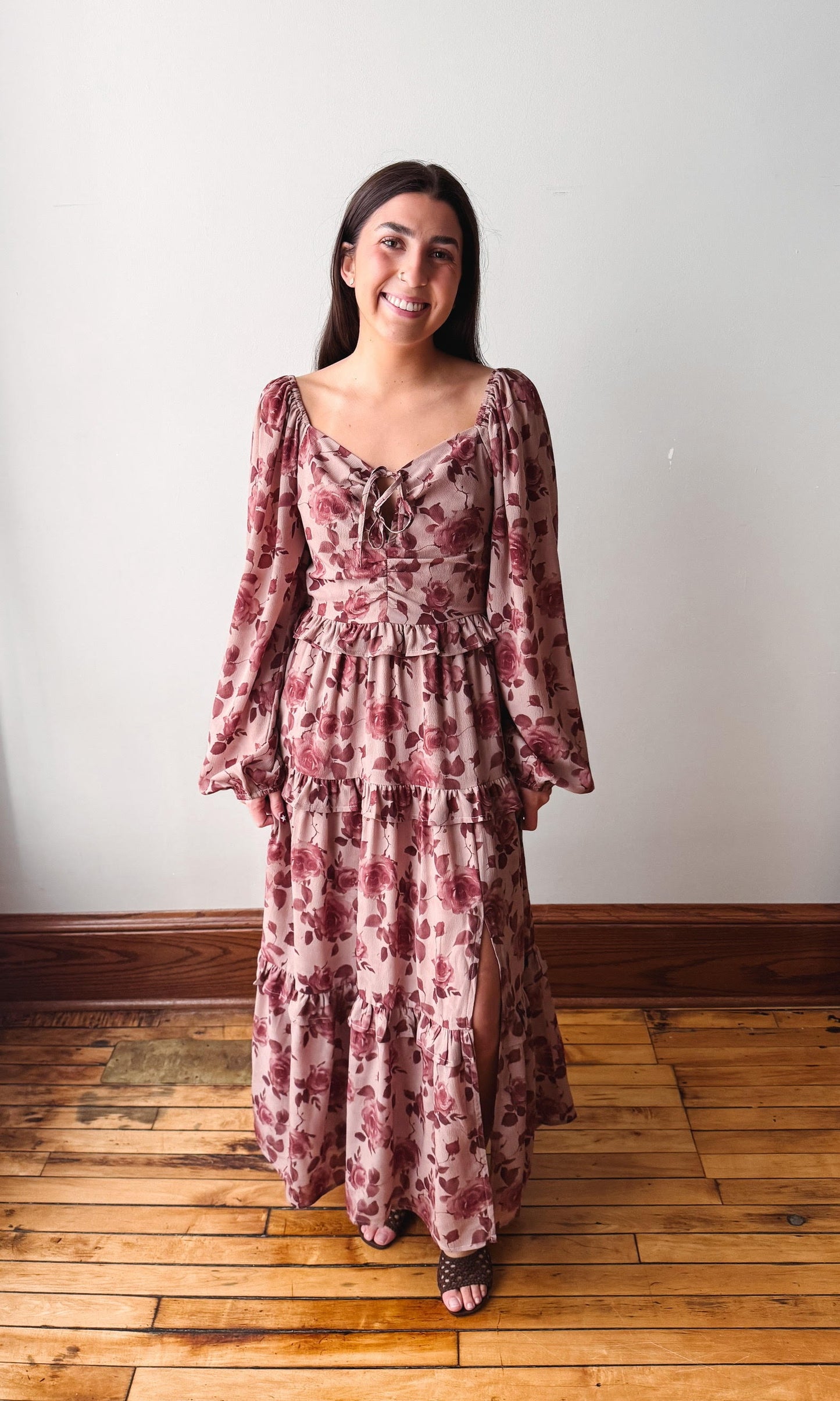 A woman wearing a long-sleeved, tiered maxi dress with a dusty rose and burgundy floral print, standing on a wooden floor against a plain off-white wall.