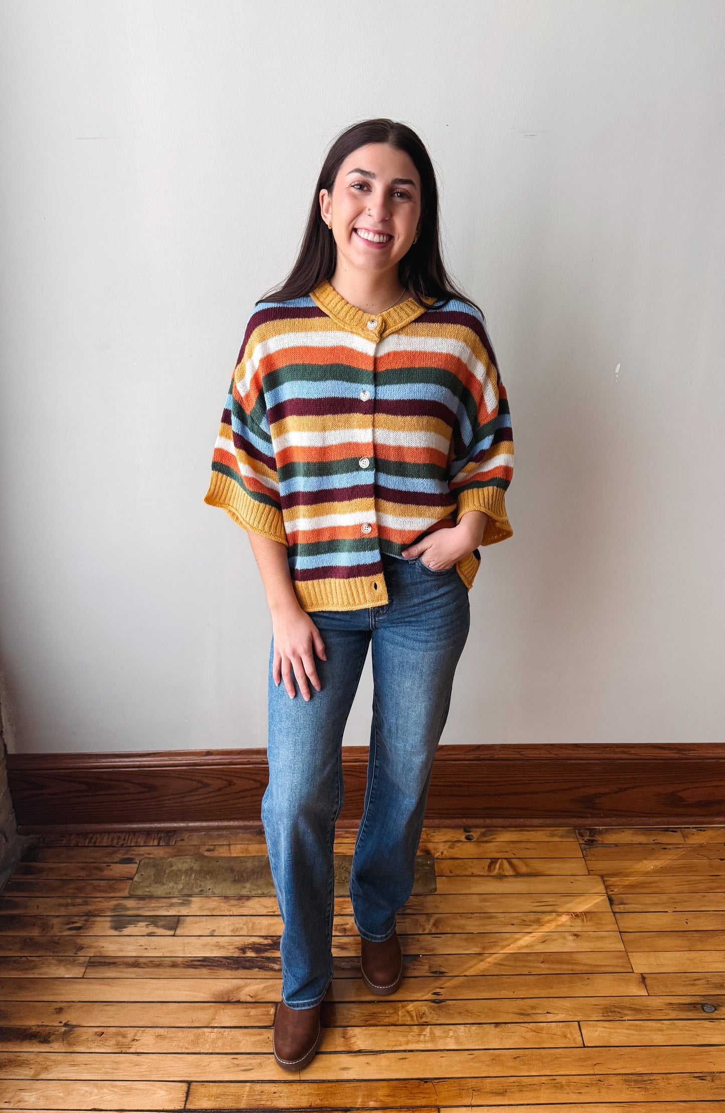 A woman stands in a room with a light gray wall and wooden floor, wearing a colorful striped knit cardigan with blue jeans and brown boots.