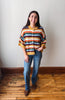 A woman stands in a room with a light gray wall and wooden floor, wearing a colorful striped knit cardigan with blue jeans and brown boots.
