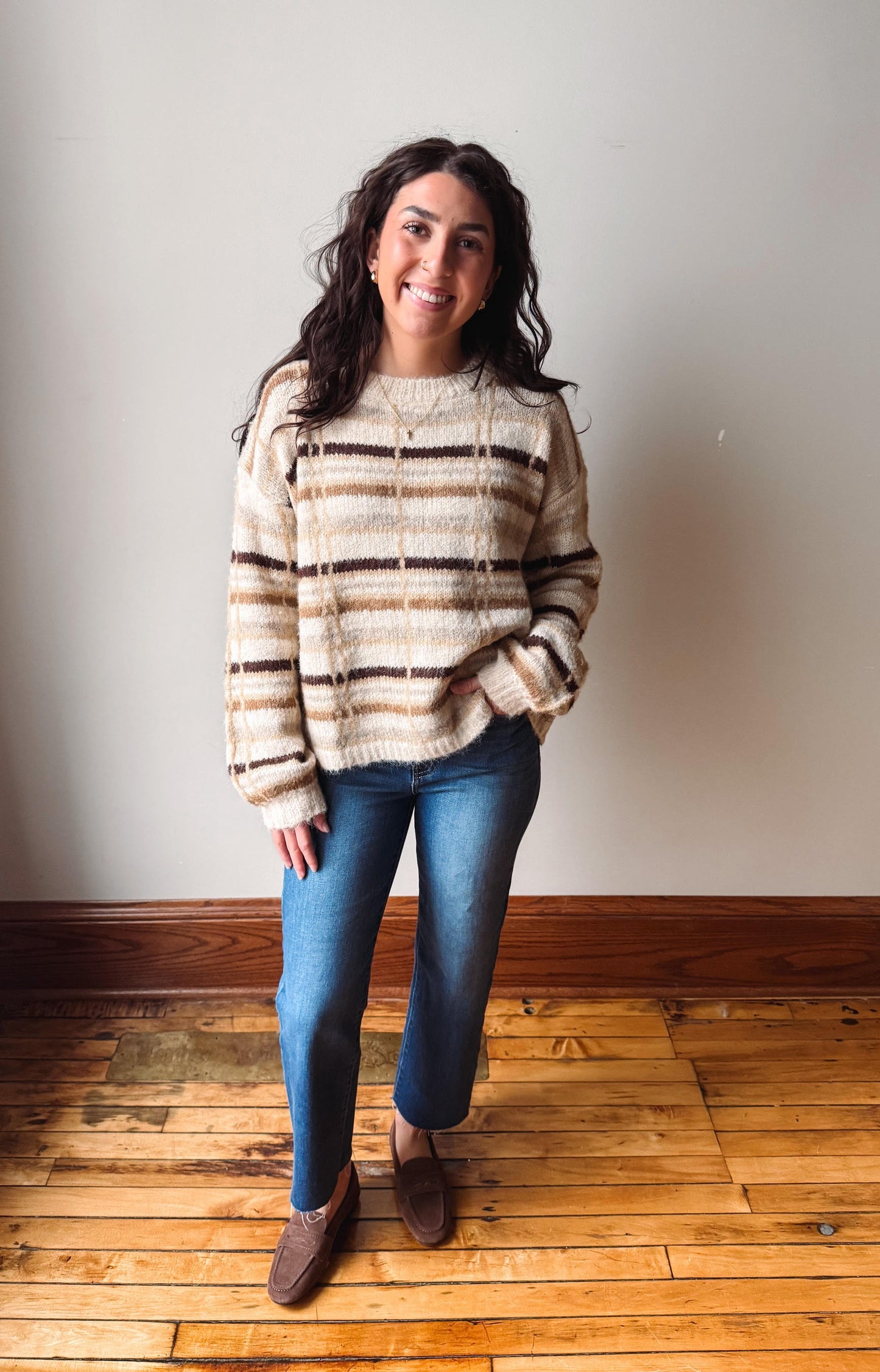 A woman stands in a room with a beige wall and wooden floor, wearing a cream and brown striped fuzzy knit sweater, blue jeans, and brown loafers.
