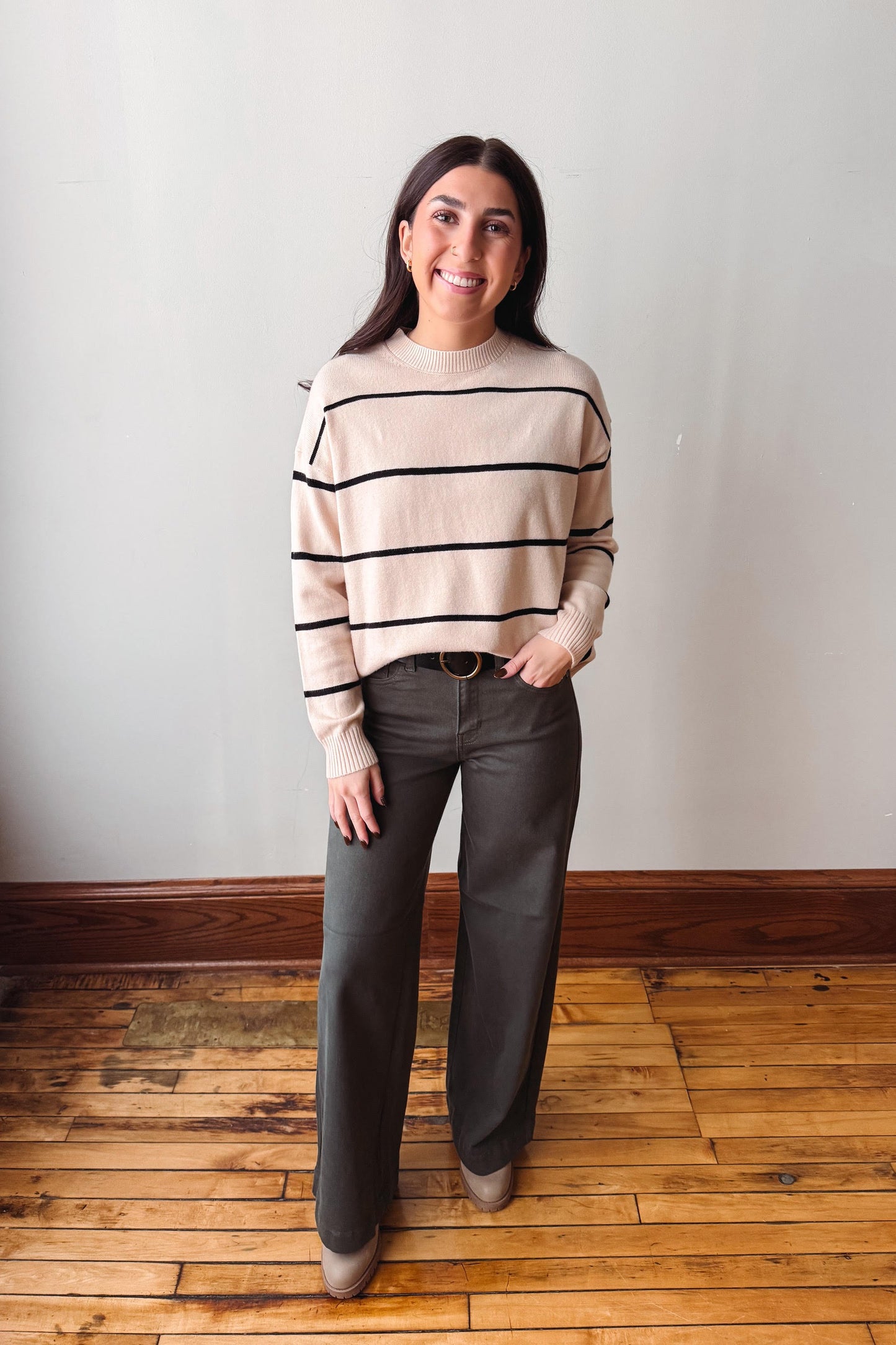 A woman stands in a room with a light gray wall and wooden floor, wearing a beige knit sweater with black horizontal stripes, dark gray wide-leg pants, and tan shoes.