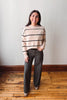A woman stands in a room with a light gray wall and wooden floor, wearing a beige knit sweater with black horizontal stripes, dark gray wide-leg pants, and tan shoes.