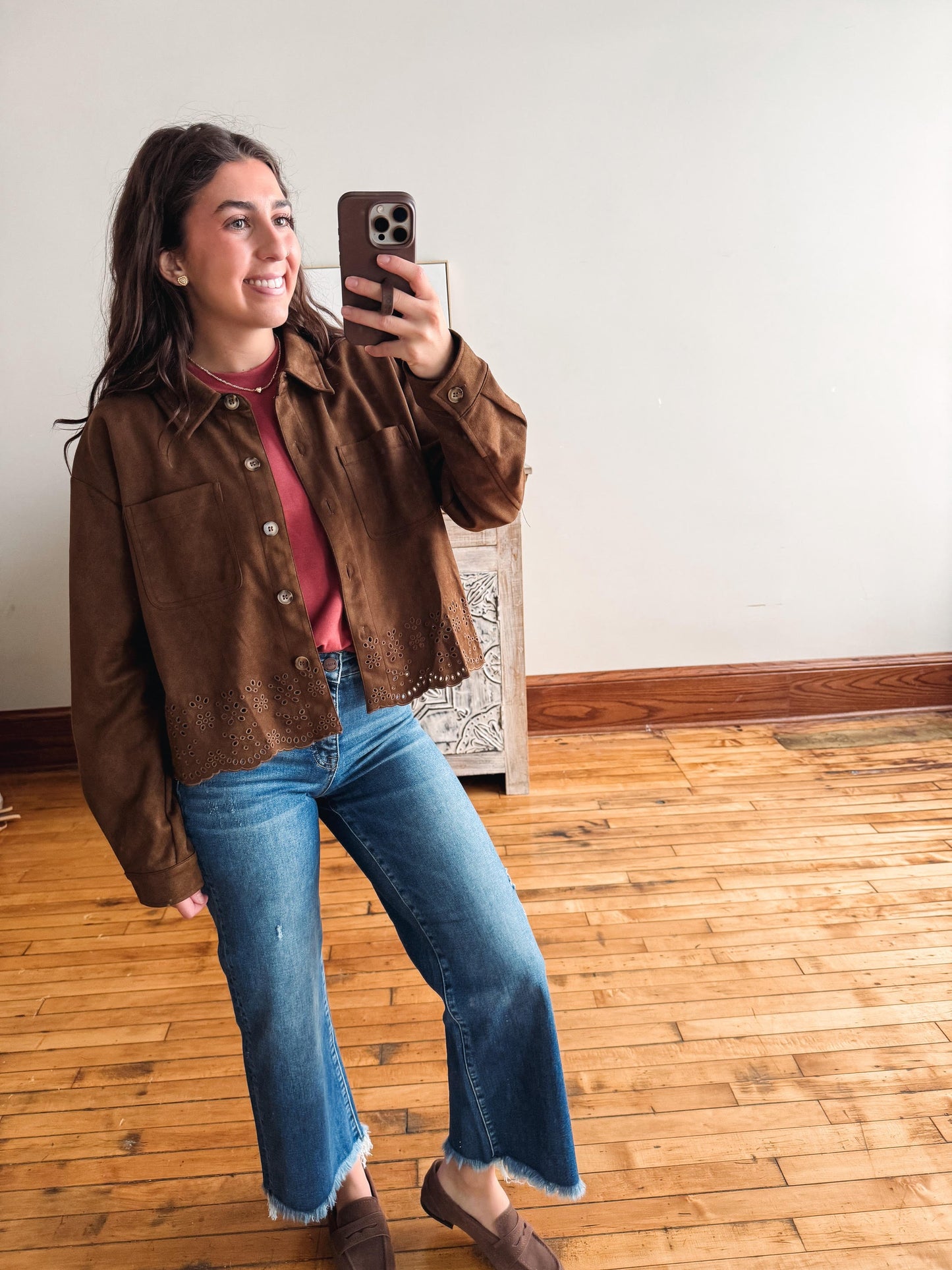 A woman takes a mirror selfie wearing a brown suede button-up shirt jacket over a dusty rose top, blue flare jeans, and brown leather loafers in a room with hardwood floors and a white wall.