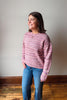 A woman wearing a pink and purple marled knit sweater and blue jeans, standing in a room with a light gray wall and wooden floor.