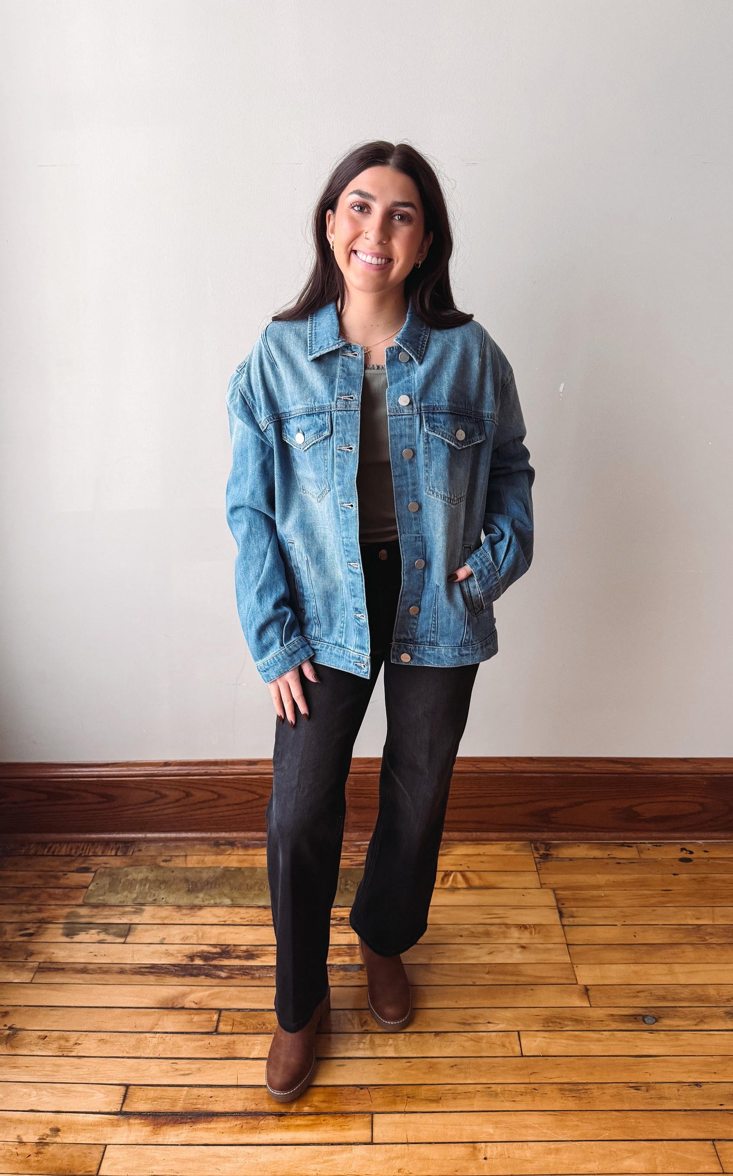 A woman stands on a wooden floor against a plain off-white wall, wearing a light blue denim jacket over a dark top and dark wide-leg pants with brown boots.