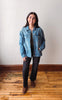 A woman stands on a wooden floor against a plain off-white wall, wearing a light blue denim jacket over a dark top and dark wide-leg pants with brown boots.