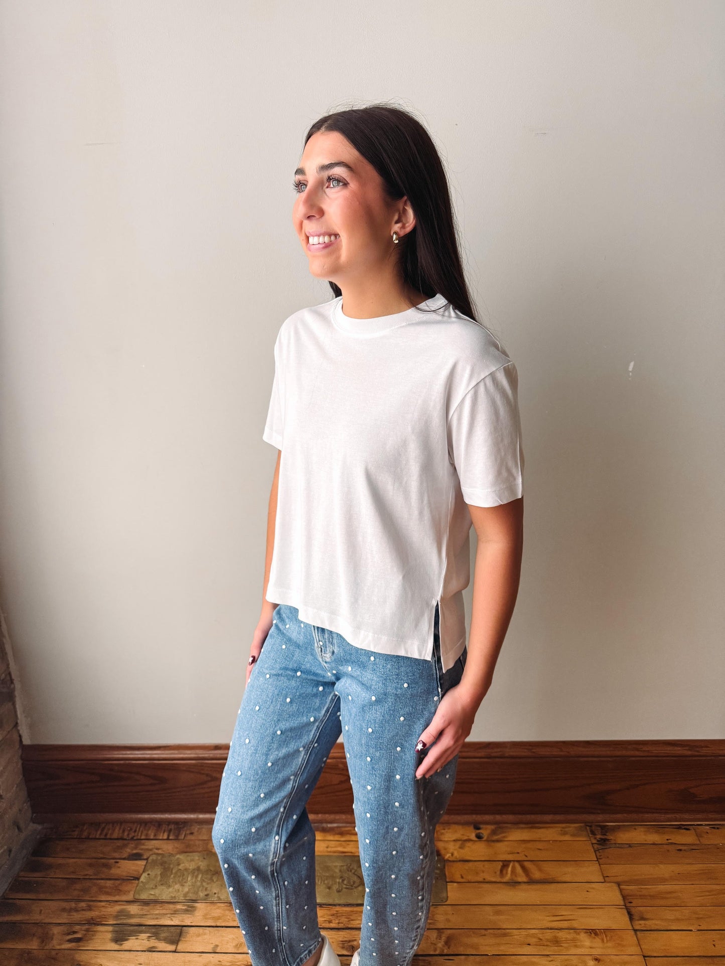 A woman wearing a white short-sleeve crewneck t-shirt and light blue jeans with small white polka dots, standing in a room with a beige wall and wooden floor.