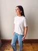 A woman wearing a white short-sleeve crewneck t-shirt and light blue jeans with small white polka dots, standing in a room with a beige wall and wooden floor.