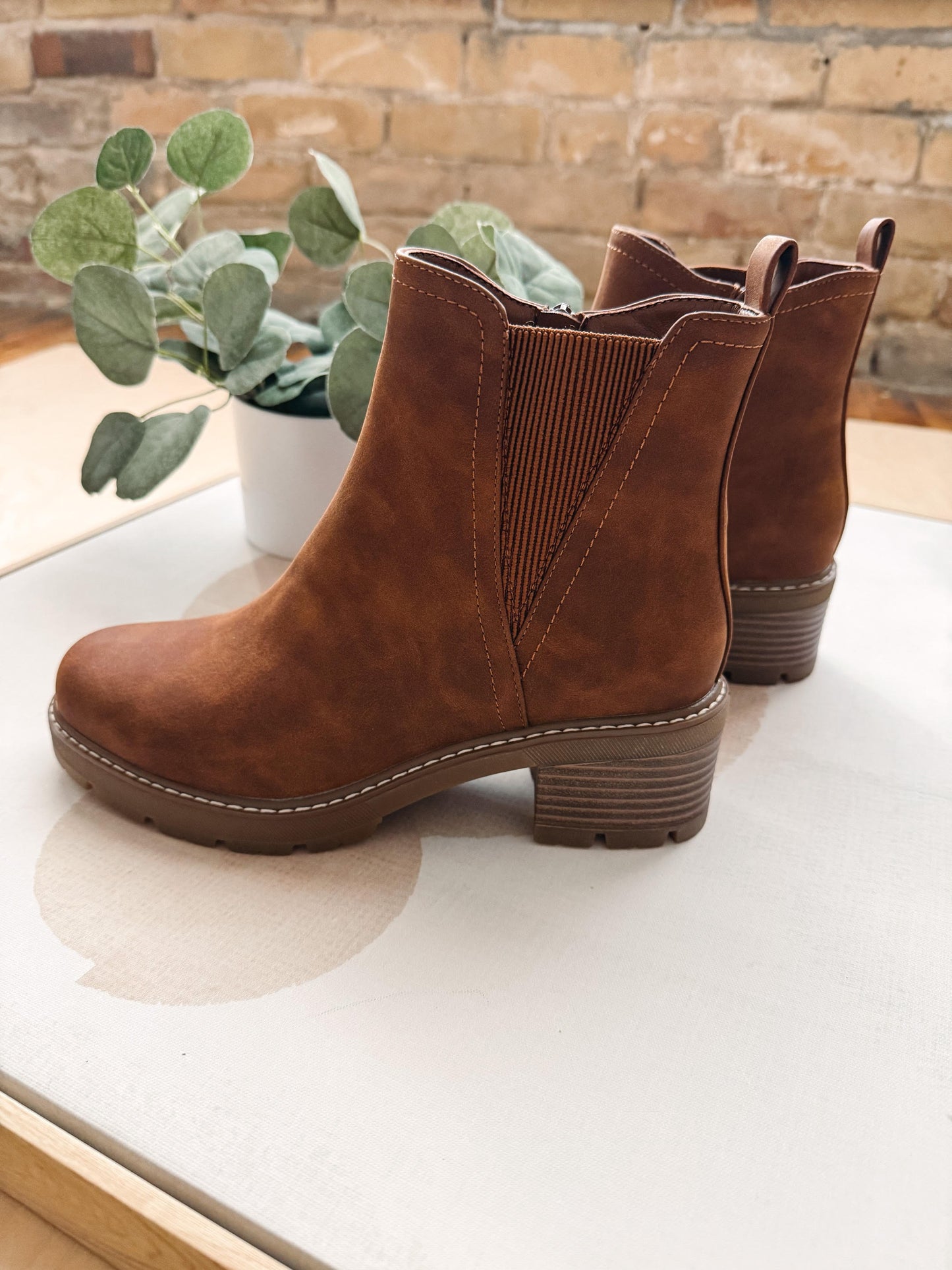 A pair of brown suede ankle boots with elastic side panels and stacked block heels, displayed on a white tabletop with a potted eucalyptus plant and a brick wall in the background.