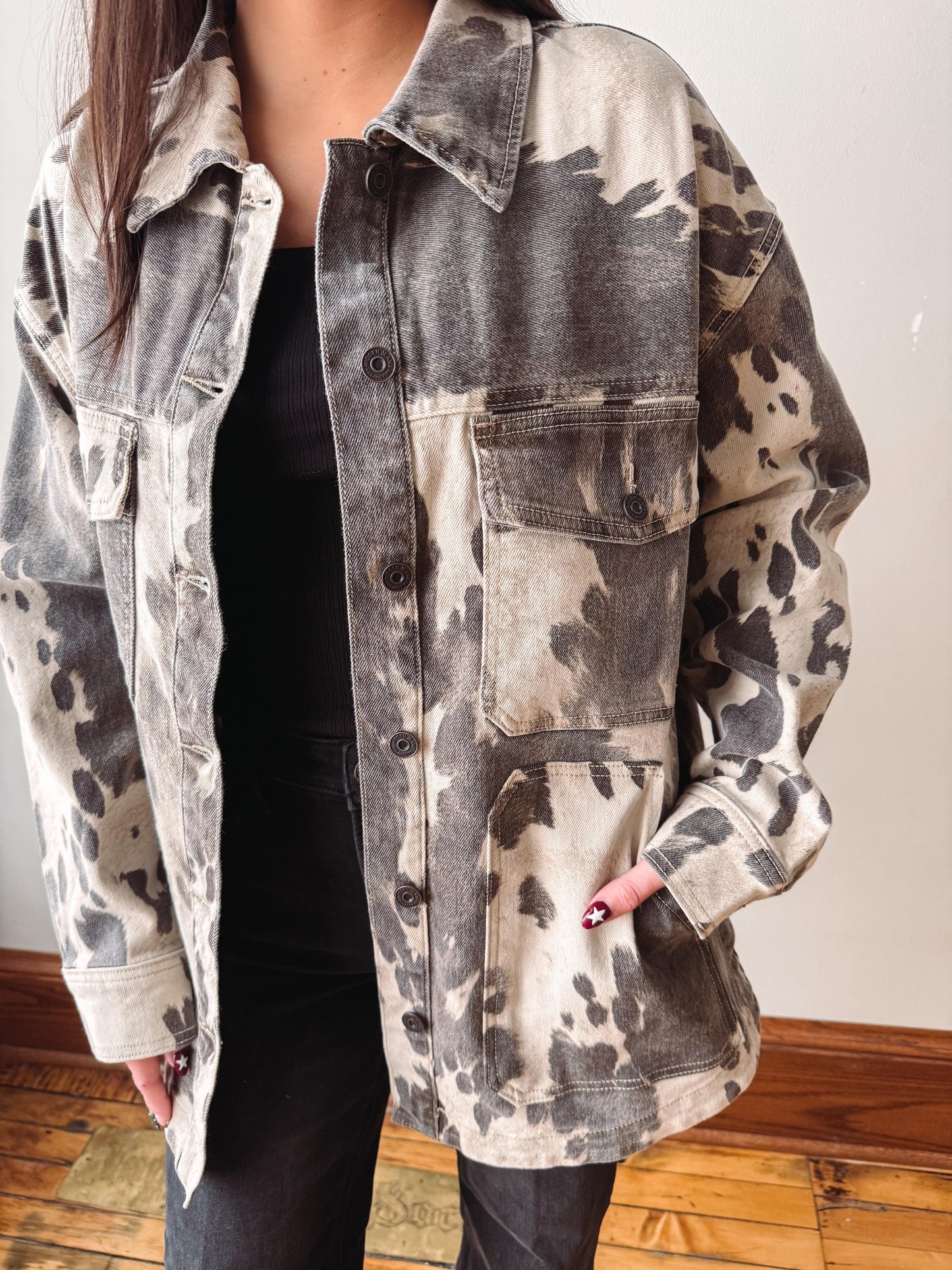 A person wearing a long-sleeve button-front overshirt with a gray and beige tie-dye or mineral-wash pattern, layered over a black top and black pants, standing indoors on a wood floor against a light wall.