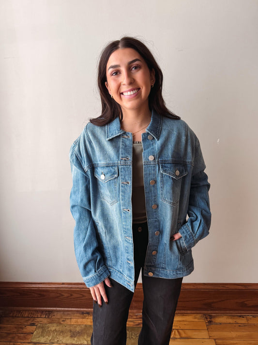 A woman smiling while wearing a light blue denim jacket over a gray top and black pants, standing against a plain off-white wall with a wooden floor visible.