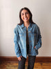 A woman smiling while wearing a light blue denim jacket over a gray top and black pants, standing against a plain off-white wall with a wooden floor visible.