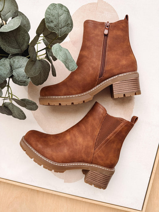 A pair of brown suede ankle boots with side zippers and chunky lug soles, arranged on a light surface with a decorative eucalyptus branch.