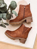 A pair of brown suede ankle boots with side zippers and chunky lug soles, arranged on a light surface with a decorative eucalyptus branch.
