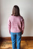 A person viewed from behind wearing a pink marled knit sweater and blue jeans, standing on a wooden floor against a plain off-white wall.