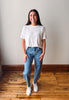 A woman stands in a room with a light gray wall and wooden floor, wearing a white short-sleeve t-shirt, blue jeans with small white star prints, and white sneakers.