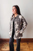 A woman modeling a gray and beige watercolor camouflage utility jacket over a black top and black jeans, standing in a room with a light gray wall and wooden floor.