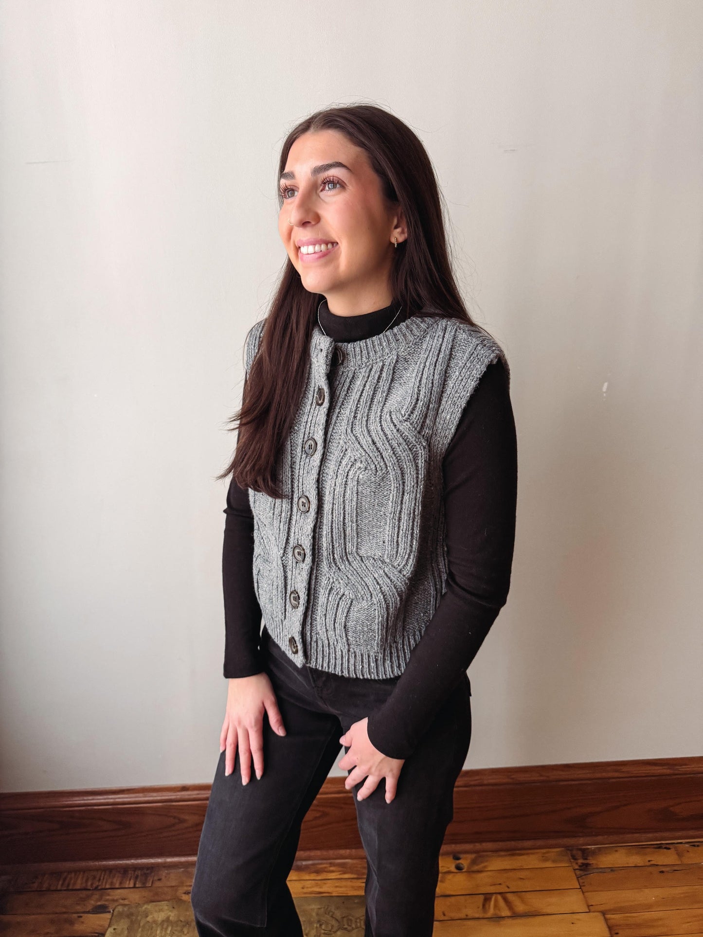 A woman wearing a gray cable-knit button-front vest over a black long-sleeve top and black pants, standing in an indoor room with a plain light wall and wood floor.