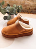 A pair of tan suede clogs with white sherpa lining and a brown platform sole, styled on a light surface with a brick wall and eucalyptus plant in the background.